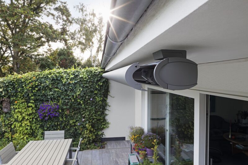 Een moderne markies in grijs, gemonteerd aan een lichte gevel met een groene heg op de achtergrond.