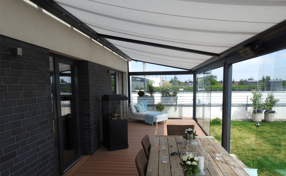 Moderne terrassensituatie met overkapping, hout en glas. Lichtgekleurde zonnescherm en meubels zichtbaar.