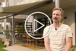 Ein Mann sitzt vor einer Terrassensituation mit Überdachung und Pflanzen im Hintergrund.