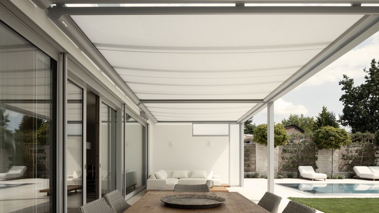 Terrassensituation mit einer weißen Markise über einem großen Holztisch, im Hintergrund ein Pool und grüne Pflanzen.