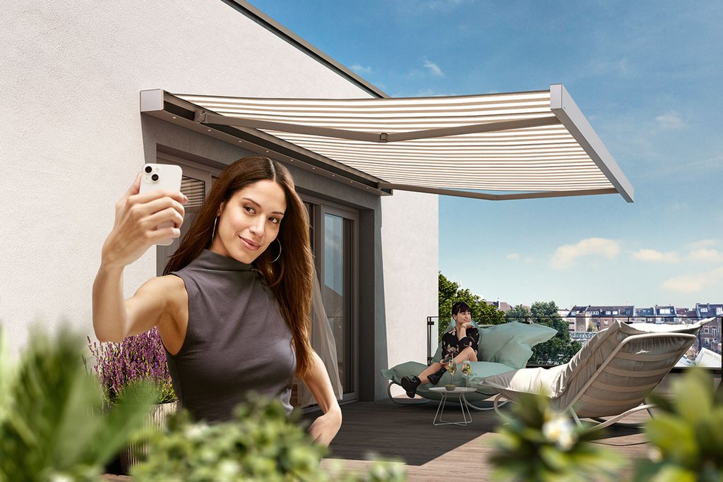 Een vrouw maakt een selfie voor een huis met een markies en een zonovergoten terras.