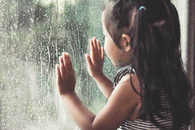 Mädchen betrachtet den Regen durch ein beschlagenes Fenster, Hände an die Scheibe gedrückt.