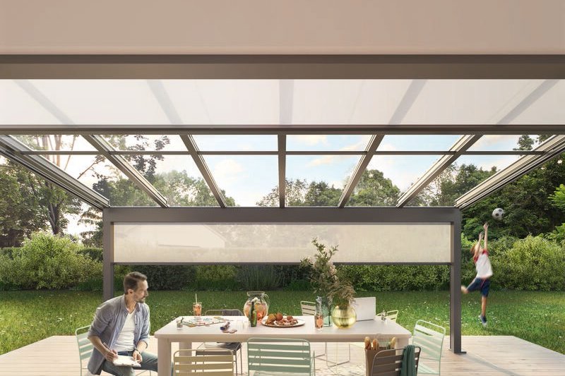 Tafel met stoelen onder een overkapping, groene tuin op de achtergrond en een kind dat speelt. Tafel met stoelen onder een overkapping, groene tuin op de achtergrond en een kind dat speelt.