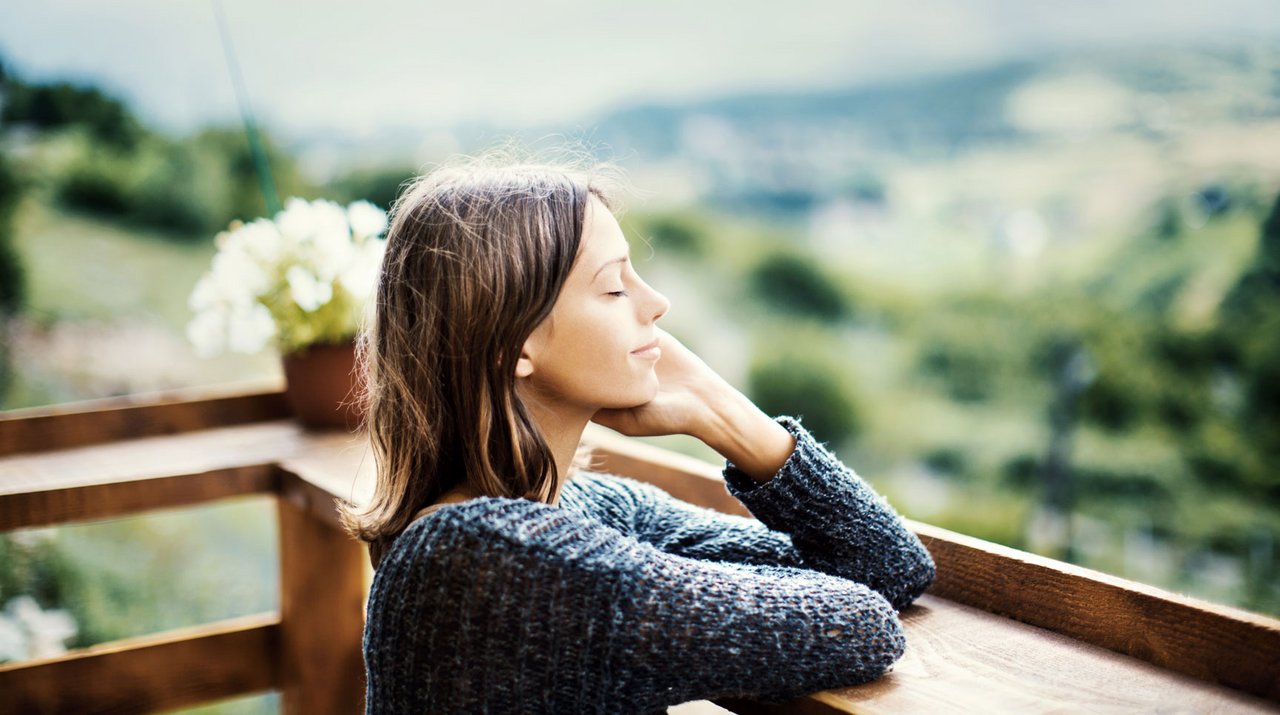 Frau mit geschlossenen Augen, die entspannt auf einer Holzterrasse sitzt, im Hintergrund grüne Landschaft.