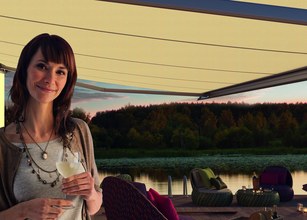 Een vrouw met een drankje onder een gele zonnescherm bij een waterlandschap. 