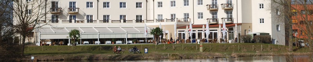 Een lichtgekleurde, moderne gebouw met een terras en groen gras aan een waterkant.
